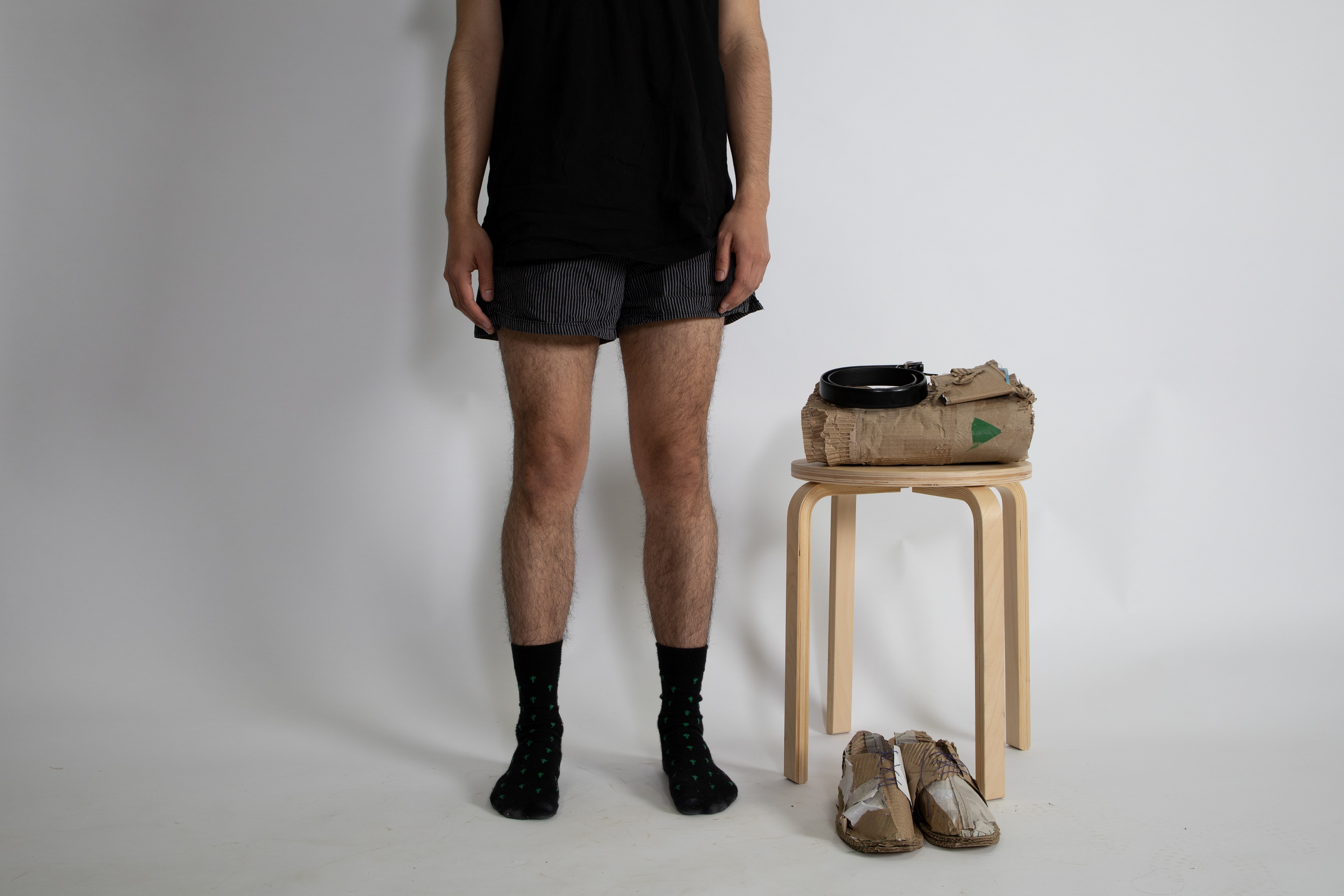 a person stnding in black socks and boxers. To the right is a short stool with folded cardboard and a belt. Bellow the stool is a pair of shoes made from cardboard